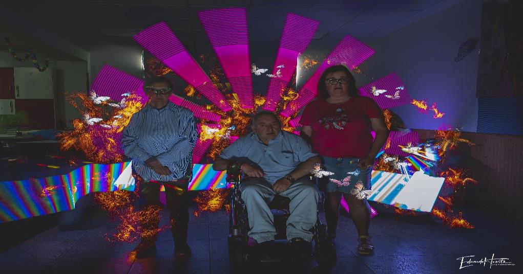 Tres participantes del CAI ASPAYM posan durante un taller de pintura con luz con efectos luminosos de colores.